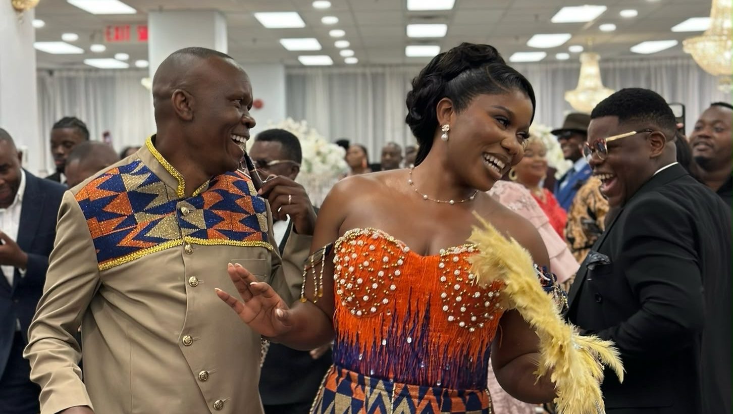 A woman in a kente mermaid gown and a man in a beige outfit with a kente bib dancing and laughing at a ceremony